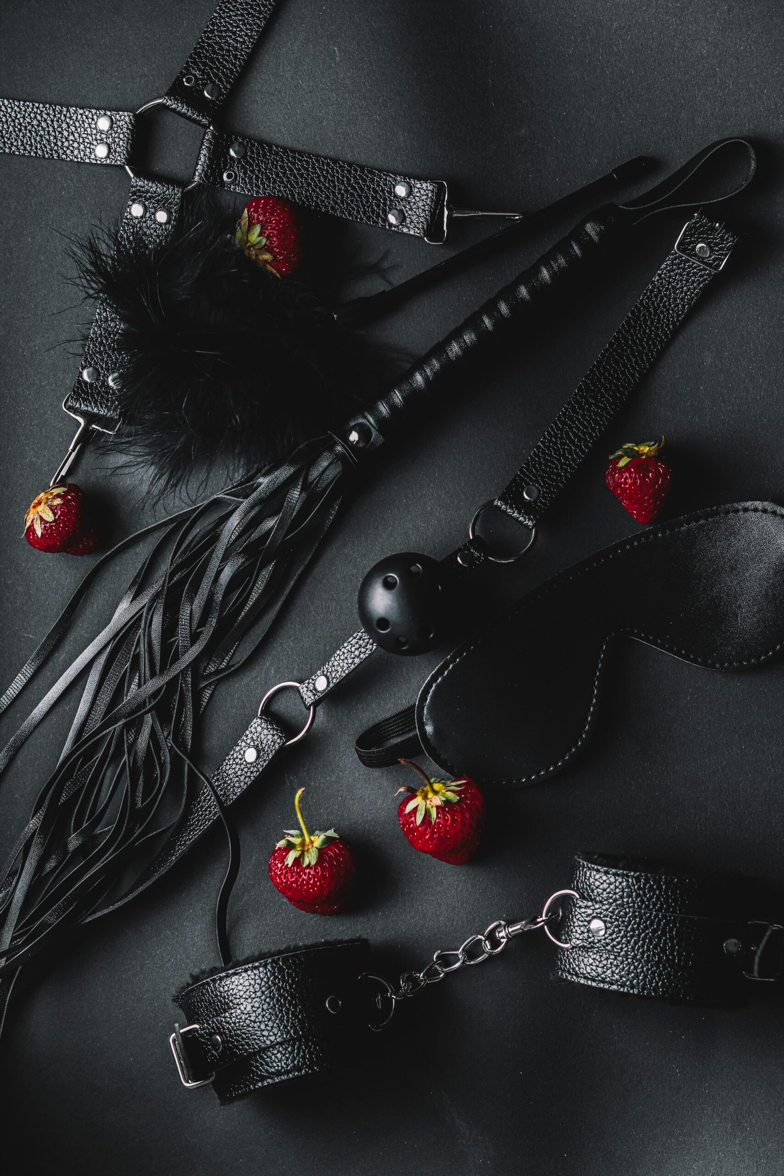 A stylish flat lay of leather BDSM accessories with fresh strawberries on a dark background.